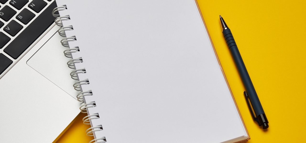 Workplace - laptop, notebook and pen for remote work on a yellow background table. Freelance desktop for home or office. Background with copy space.